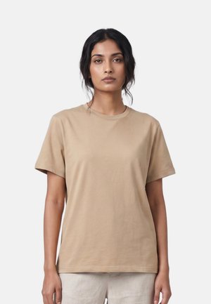 Vrouw met donker haar draagt een effen beige T-shirt met korte mouwen en lichte broek, staand tegen een neutrale lichte achtergrond.