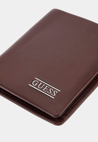 Portafoglio in pelle marrone con una superficie liscia, caratterizzato da un logo "GUESS" argentato sulla parte anteriore e bordi cuciti. Design compatto.