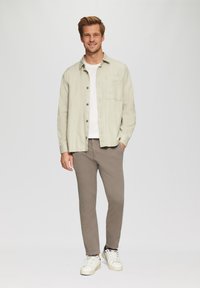 Chemise boutonnée vert clair sur un t-shirt blanc, assortie à un pantalon gris et des baskets blanches. La chemise a une poche poitrine.