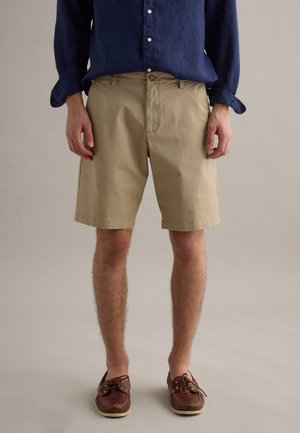 Mann trägt beigefarbene knielange Shorts, marineblaues Langarmshirt und braune Leder-Bootsschuhe, steht vor neutralem Hintergrund.
