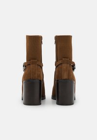 Botine din suede maro, cu partea superioară texturată, fermoar lateral și toc lat. Prezintă o curea cu o cataramă metalică ca accent.