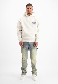 Lichtgrijze hoodie met een voorzak en logodetail, gecombineerd met vervaagde blauwe spijkerbroek en witte sneakers.
