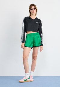 Black cropped long-sleeve shirt with white stripes, green satin shorts, pink sneakers with green accents, and round sunglasses.