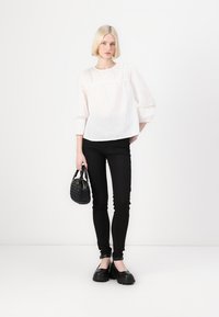 Bílá, strukturovaná blůza s krajkovými detaily, přiléhavé černé kalhoty a masivní černé boty. Modelka drží malou černou kabelku.