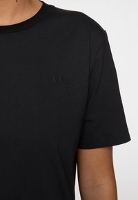 Musta T-paita pehmeästä kankaasta, jossa on pyöreä kaula-aukko. Siinä on huomaamaton musta logo vasemmassa rinnassa ja lyhyet hihat.