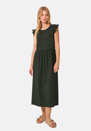 Femme portant une robe midi noire sans manches avec des épaules à volants et des sandales beiges à bout ouvert, debout devant un fond uni.