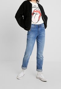 Černá chlupatá bomber bunda přes bílý tričko s grafikou Rolling Stones, spárovaná se světle modrými př rolled-up džíny a bílými teniskami.
