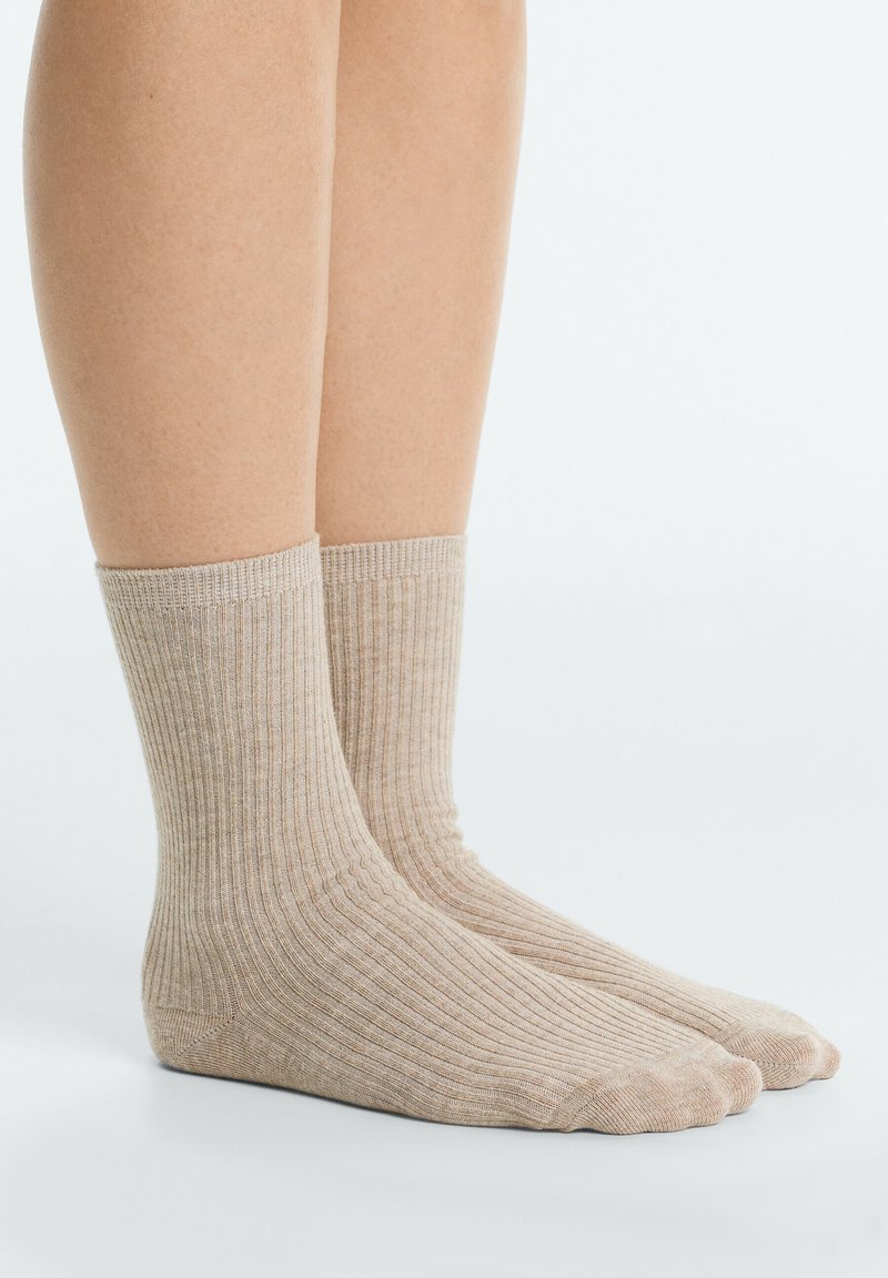 Paire de pieds portant des chaussettes côtelées beige mi-mollet, debout sur une surface blanche sur un fond uni.