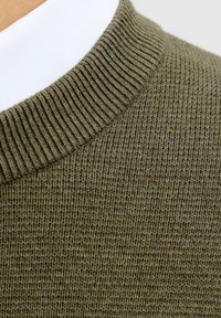 Tuvumā redzams ribots olīvzaļš adīts džemperis ar apkakli, kas uzlikta virs baltas kreklu.