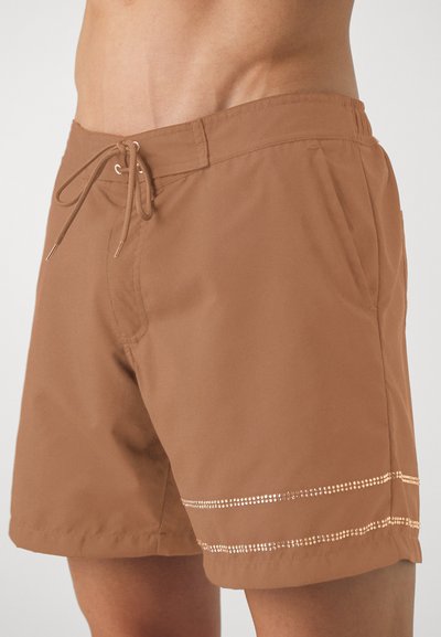 Brune badedraktshorts laget av lett materiale, med snor i midjen, sidelommer og dekorative metallisk detaljer nær nederkanten.