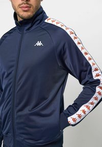 Jachetă de trening bleumarin cu guler înalt, închidere cu fermoar și bandă cu imprimeu logo alb și portocaliu în contrast pe mâneci.
