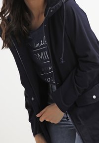 Mörkblå zip-up jacka med snoddhuva, med sidofickor och silverfärgade knappar. Bärs över en mörk grafisk t-shirt med vit text.