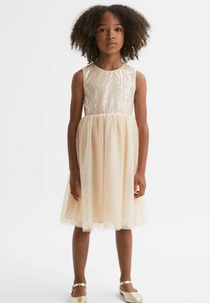 Młoda dziewczyna o kręconych włosach w beżowej sukience bez rękawów z cekinowym gorsetem i tiulową spódnicą, stojąca na jednolitym białym tle.