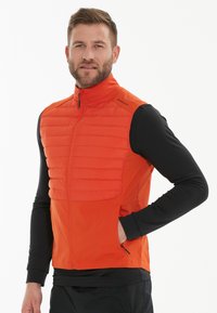 Orange isolerad väst med kviltad front, dragkedja och hög krage. Bärs över en svart långärmad skjorta med sidofickor.