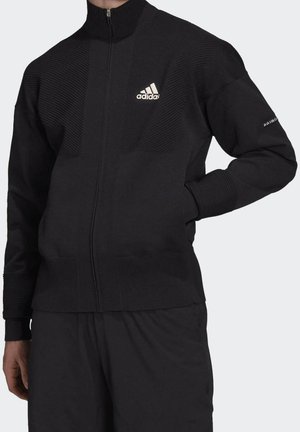 Zwarte Adidas jas met volledige ritssluiting, geribbelde structuur, logo op de borst en tekst "prime" op de linker mouw, gedragen met bijpassende zwarte broek.
