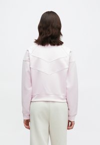 Άτομο με καστανά μαλλιά μήκους μέχρι τον ώμο, φορώντας ροζ φούτερ και λευκά παντελόνια, στέκεται απέναντι σε άσπρο φόντο.