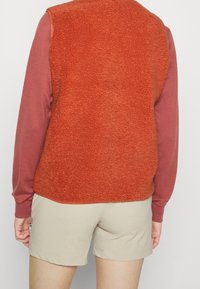 Gilet in pile color arancio bruciato, con texture morbida, colletto alto e design senza maniche, abbinato a una camicia a maniche lunghe color vino e pantaloni corti beige.