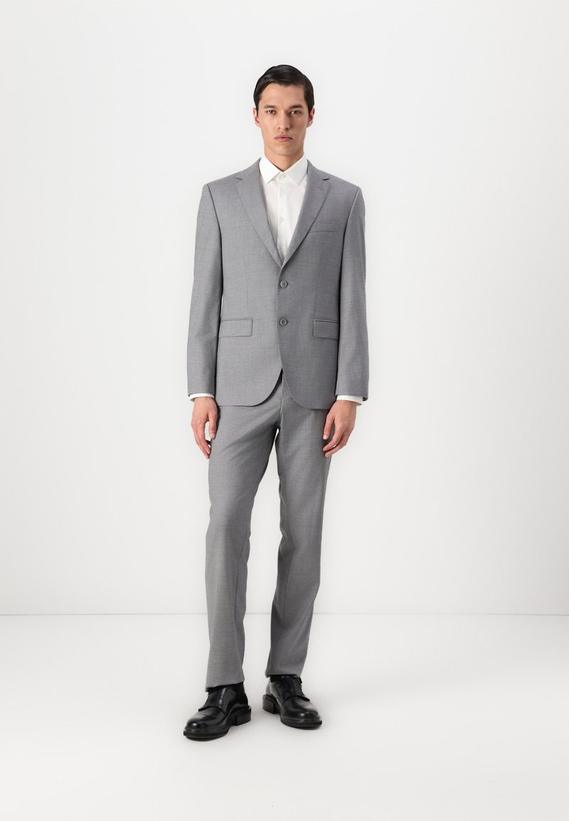 Completo grigio con rever a punta, due bottoni e tasche a pattina. Abbinato a una camicia bianca. Indossato con scarpe in pelle nere. Texture liscia.