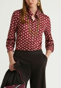 Maroon button-up shirt featuring a geometric pattern in cream and gold. Long sleeves with rounded cuffs. Smooth, shiny fabric texture.