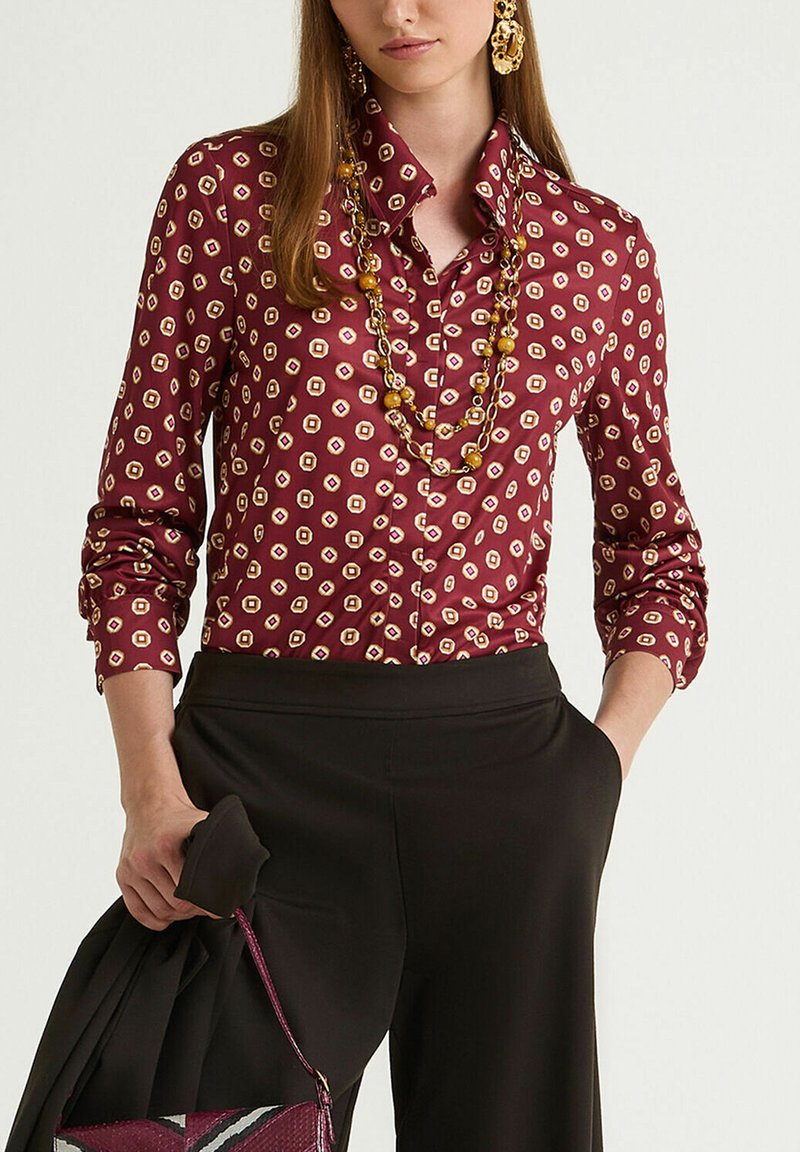 Maroon button-up shirt featuring a geometric pattern in cream and gold. Long sleeves with rounded cuffs. Smooth, shiny fabric texture.