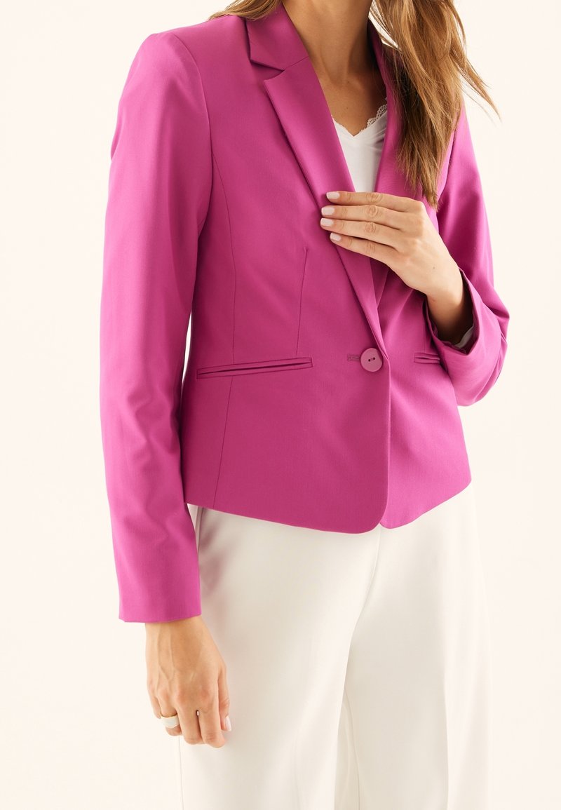 Fuksjowa marynarka z wyciętym klapem, zapinana na jeden guzik, z dwoma przednimi kieszeniami, zestawiona z kremowymi spodniami. Gładka faktura tkaniny.