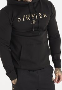 Hombre vestido con sudadera negra con el texto beige "SIKSILK" y pantalones de chándal negros, con tatuajes visibles en sus manos, de pie frente a un fondo blanco.
