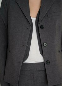 Grijze op maat gemaakte blazer met zwarte knopen, één pocket en een gladde textuur, gecombineerd met een witte aansluitende top.