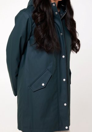Femme portant un manteau imperméable à capuche bleu sarcelle foncé avec des boutons-pression et des poches à rabat, visible de la poitrine à mi-cuisse.