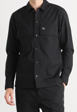 Mann trägt ein schwarzes, langärmeliges Hemd mit Brusttaschen und einem dezenten „ck“-Logo auf der linken Tasche.