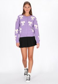 Liliowy sweter z wzorem białych kokardek, zapinany z przodu, z prążkowanymi mankietami i dołem. W zestawie z czarną plisowaną spódnicą i czarnymi sneakersami.