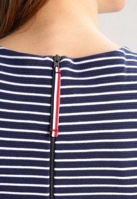 Gros plan du dos d'une chemise rayée bleu marine et blanche avec une fermeture éclair noire et une languette en tissu rouge, blanche et bleue près du col.