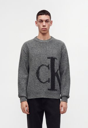 Joven con un suéter gris de punto con un gran logo negro de "CK", de pie frente a un fondo blanco liso.