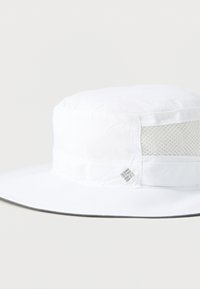 Λευκό καπέλο τύπου bucket με στρογγυλό, επίπεδο στέμμα και φαρδύ γείσο. Πάνελ από δίχτυ στις πλευρές για αερισμό. Μικρή λεπτομέρεια με ραμμένο λογότυπο στο μπροστινό μέρος.