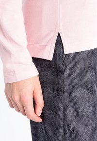 Jasnoróżowa bluzka z długim rękawem o fakturowanej tkaninie, z bocznymi rozcięciami. W połączeniu z ciemnoszarymi spodniami w wzory. Dłoń spoczywa na tkaninie.