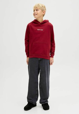 Sudadera roja con un bolsillo frontal y un logotipo blanco bordado, combinada con pantalones oscuros gris sueltos y zapatos negros.