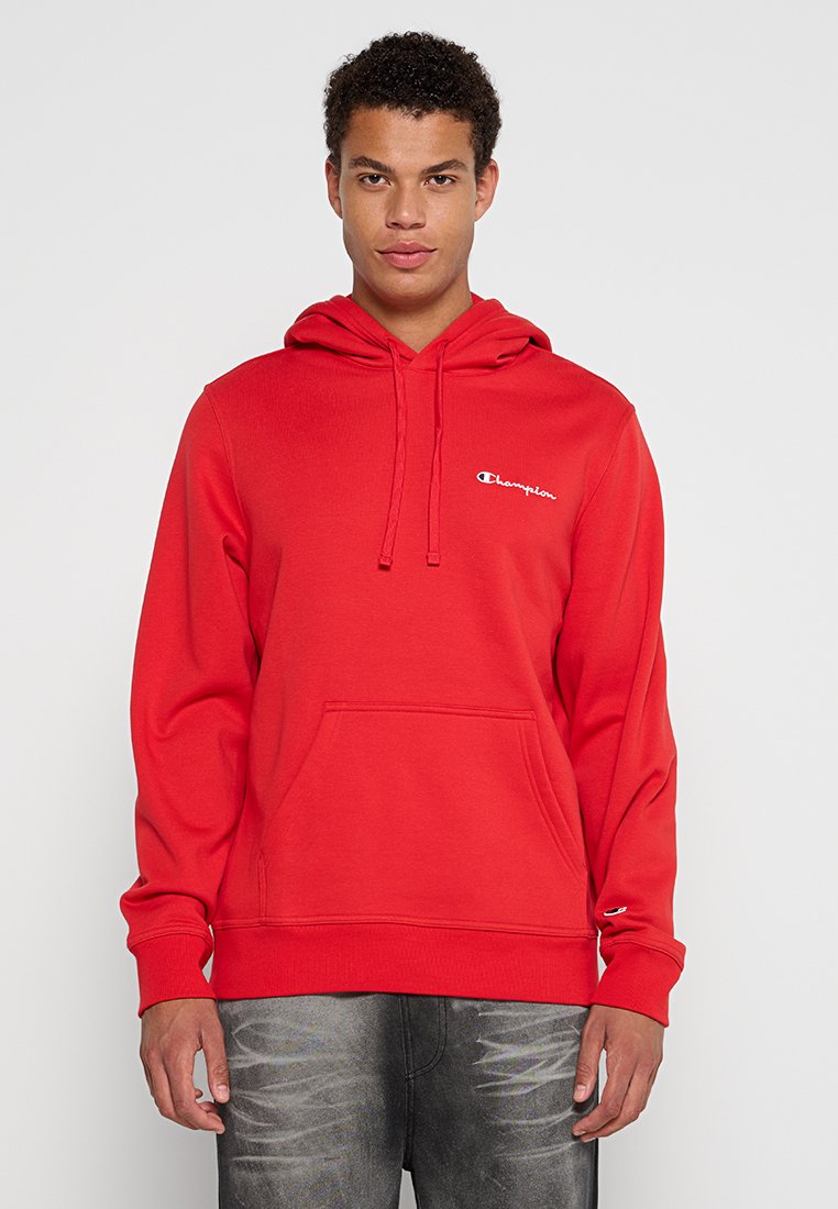 Joven vistiendo una sudadera roja de Champion con bolsillo frontal y cordones, combinada con vaqueros gris oscuro frente a un fondo liso.