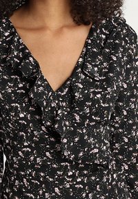 Musta kukkamekko, jossa on V-pääntie ja röyhelödetalji, pienet vaaleanpunaiset kukat ja vihreät varret halkovat kangasta.