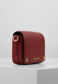 Borsa a tracolla rossa in pelle con finitura testurizzata, dettagli in oro e pattina pieghevole. Presenta un logo sulla parte anteriore e una tracolla decorativa.