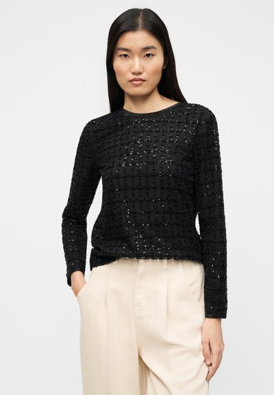 Μαύρο μακρυμάνικο τοπ με υφασμένο μοτίβο πλέγματος και διακοσμητικά στοιχεία. Σχεδιασμένο με στρογγυλή λαιμόκοψη, συνδυασμένο με ανοιχτό-beige παντελόνι.