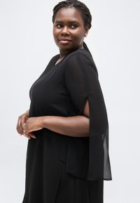 Γυναίκα με πλεξούδες, που φοράει χρυσούς κρίκους στα αυτιά και μαύρο φόρεμα με διαφανείς, σχισμένες μανίκες, κοιτάζοντας λίγο προς το πλάι σε λευκό φόντο.