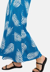 Ein blaues Stoffkleid mit weißen Blattmustern. Eine Person trägt schwarze flache Sandalen, der Fokus liegt auf dem fließenden Design des Kleides.