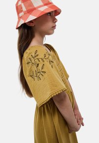 Robe en coton jaune à manches courtes avec broderie florale, associée à un chapeau seau à carreaux rouges et blancs. Texture lisse, design superposé.