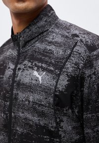 Homme portant une veste de sport noire et grise à motif avec une fermeture éclair et un logo Puma blanc sur la poitrine.