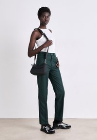 Pantalon vert sur mesure, top sans manches blanc et sac à bandoulière noir. Baskets noires avec accents blancs. Textures lisses et design structuré.
