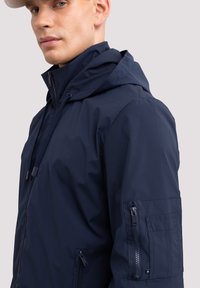 Tumed siniset kapuutsiga jakk, mis on valmistatud kergest materjalist. Jakk on varustatud lukuga ees, reguleeritava stringiga ja lukuga käepojaga.