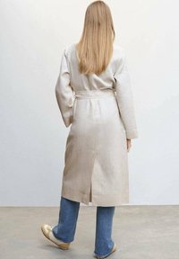 Beige linnedragt med knyt i taljen, lange ærmer og slids bagpå, parret med blå jeans og lyse sko.