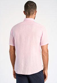 Camicia in lino rosa chiaro a maniche corte con colletto classico, tessuto strutturato e orlo curvo, indossata con pantaloni corti scuri.