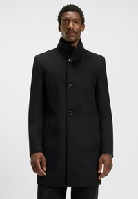 Cappotto di lana nera con colletto alto, sei bottoni e tasche laterali. Texture liscia con una vestibilità su misura. Design semplice, senza motivi o accenti.