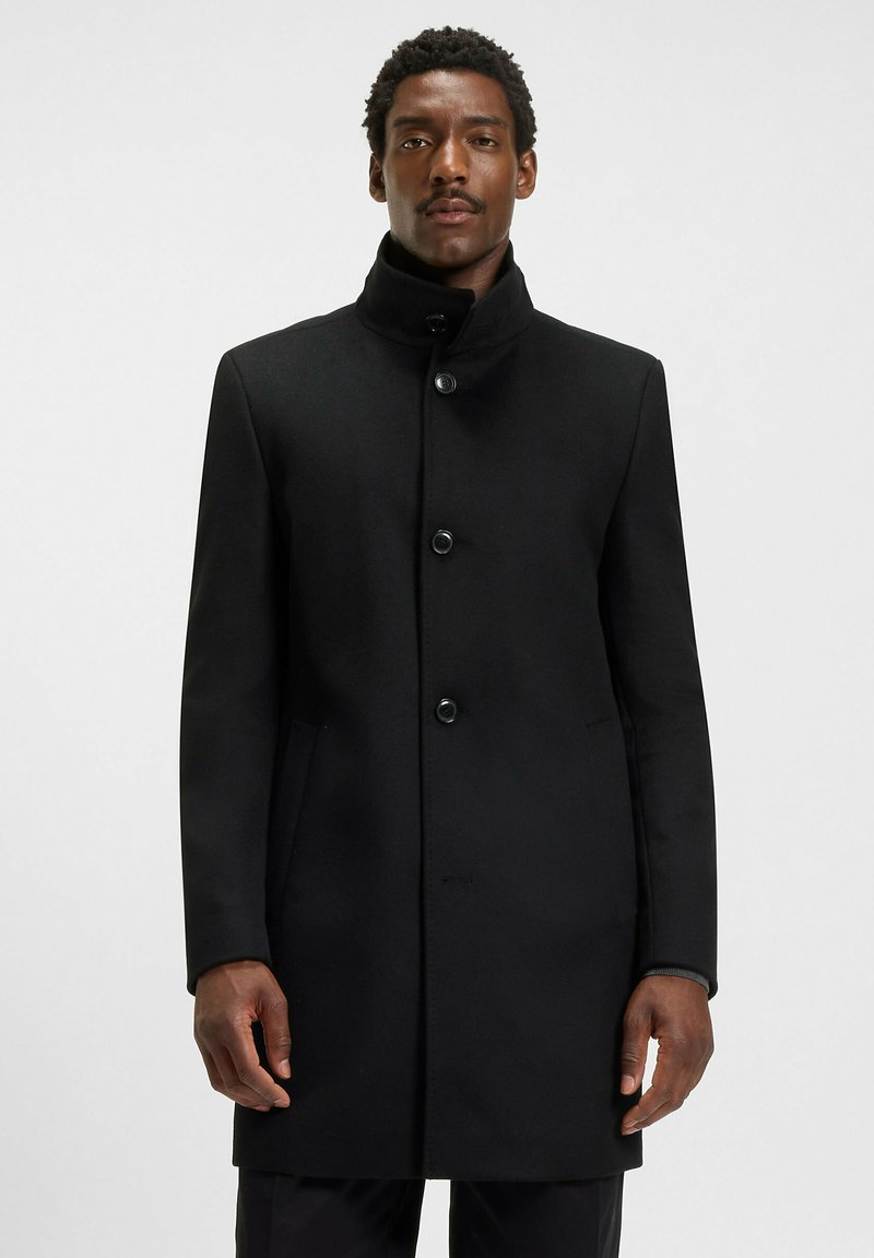 Cappotto di lana nera con colletto alto, sei bottoni e tasche laterali. Texture liscia con una vestibilità su misura. Design semplice, senza motivi o accenti.