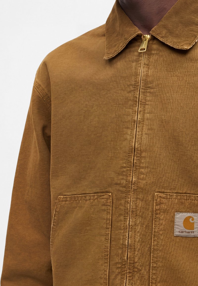 Brown zip-up jacket with two front pockets, collar, and a Carhartt logo patch on the right pocket, worn by a person.